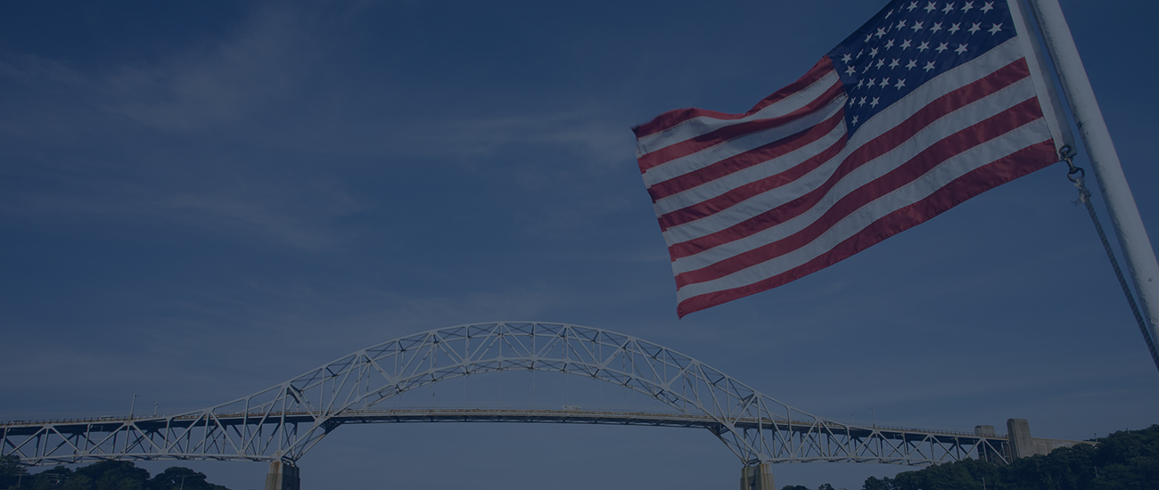 A raised flag of the United States of America with a bridge in the background.